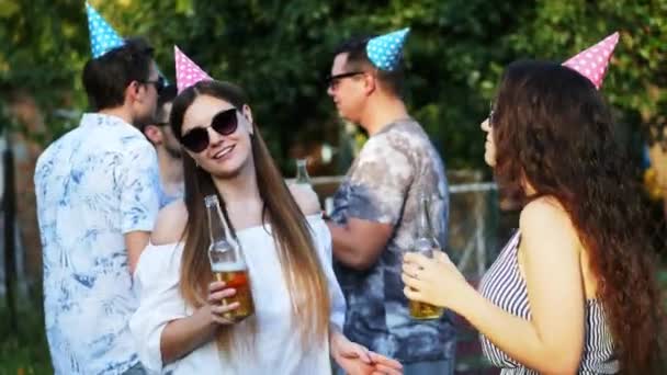 Két boldog nő beszél és iszik sört a nyári party, a férfiak a háttérben, fárasztó üdülési sapka — Stock videók