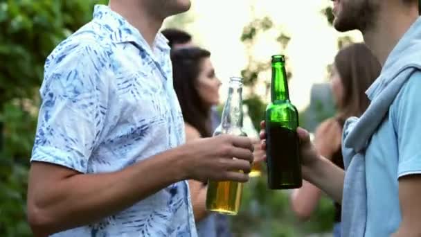 Deux amis tiennent des bouteilles de bière dans leurs mains et parlent, en arrière-plan les gens dansent à la fête — Video