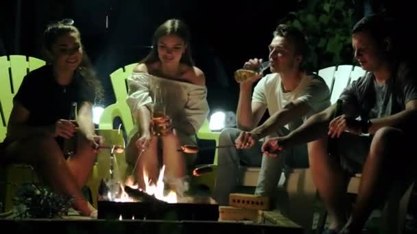 Venner som sitter ved bålet om kvelden, steker pølser, snakker og drikker øl fra flaske – stockvideo