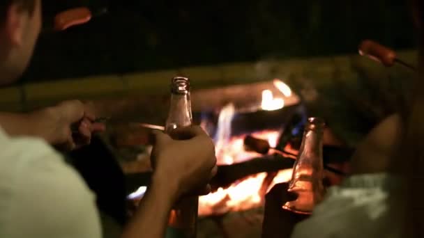 Primo piano di bottiglie di birra nelle mani di amici, salsicce su bastoncini arrosto sul fuoco sullo sfondo, un gruppo di persone che si rilassano vicino al falò — Video Stock