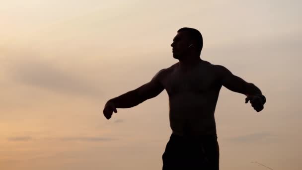 Silhueta de homem atlético com um tronco nu posando mostra seus músculos e bíceps contra o céu ao pôr do sol. Desporto, saúde e fitness — Vídeo de Stock