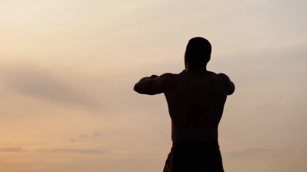 Silhueta de homem atlético com um tronco nu posando mostra seus músculos contra o céu ao pôr do sol. Desporto, saúde e fitness — Vídeo de Stock