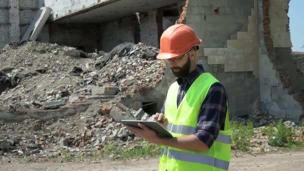 Bir kask ve yansıtıcı yelek sakallı oluşturucu yıkım için hazırlanıyor, yıkılmış bir binanın arka planda bir tablet kullanır — Stok video