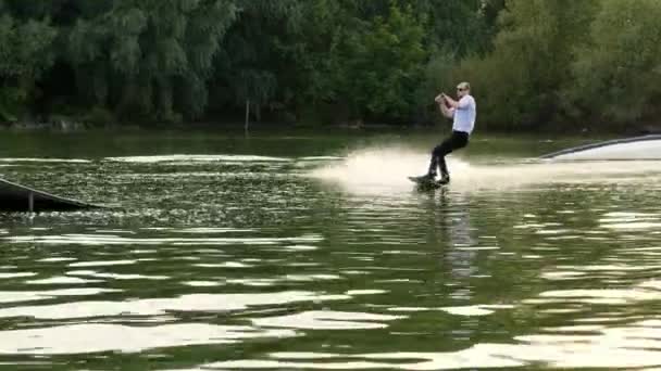 Hombre de negocios extremo en un traje con corbata y gafas de sol monta en wakeboard y saltando en un trampolín en un lago — Vídeo de stock