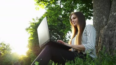 Ağacın yanındaki çimenlerin üzerinde oturan iş kadını gün batımında parkta dizüstü bilgisayar kullanıyor