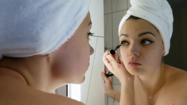 Mulher bonita em uma toalha na cabeça aplica maquiagem, pinta seus olhos e pestanas de cor na frente de um espelho — Vídeo de Stock