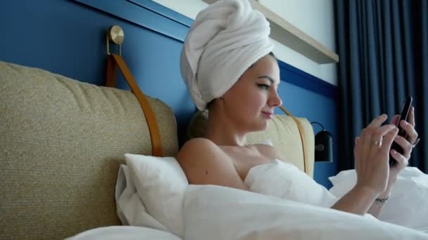 Schöne, glückliche Frau liegt nach der Dusche mit einem Handtuch auf dem Kopf im Bett und benutzt Smartphone, blättert Seiten und surft im Internet. — Stockvideo