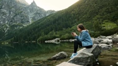Mutlu kadın gezgin bir dağ gölü üzerinde cep telefonu ile mobil oyun oynarken, doğaya dinleniyor