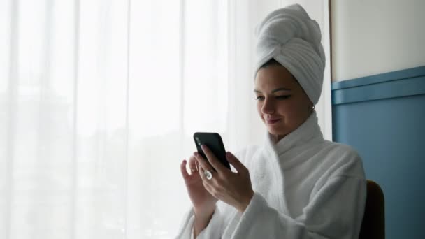 Close-up portret van vrouw in badjas, met handdoek op haar hoofd, maakt gebruik van mobiele telefoon — Stockvideo