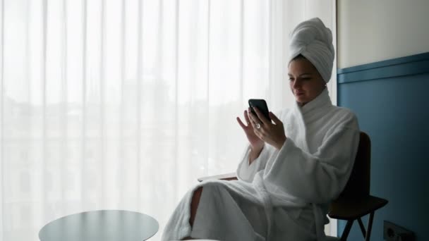 Retrato de mulher bonita em roupão de banho, com toalha na cabeça, usa telefone celular — Vídeo de Stock