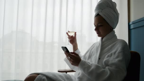 Mujer elegante en un albornoz blanco y una toalla en la cabeza disfrutando de una copa de vino y el uso de teléfono móvil en el hotel — Vídeo de stock