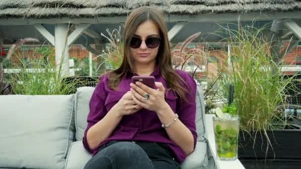 매력적인 젊은 여성은 휴대 전화를 사용하고 차가운 모히토 칵테일을 마십니다. 테라스에서 즐거운 시간을 보내고 — 비디오
