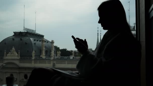 Close-up silhouette of woman in bathrobe uses mobile phone in a hotel room, beautiful museum in a large window — Stock Video