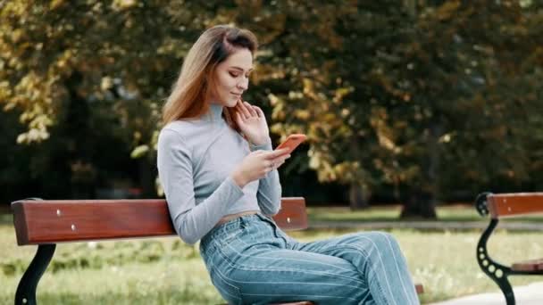 Chica pelirroja feliz estudiante usando el teléfono móvil en un banco en un parque, mujer joven sonriendo mientras usa la aplicación — Vídeo de stock