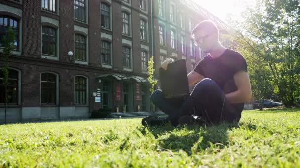 캠퍼스의 녹색 잔디밭에 있는 공원에 앉아 노트북을 작업하는 현대 남성 학생 또는 프리랜서 — 비디오