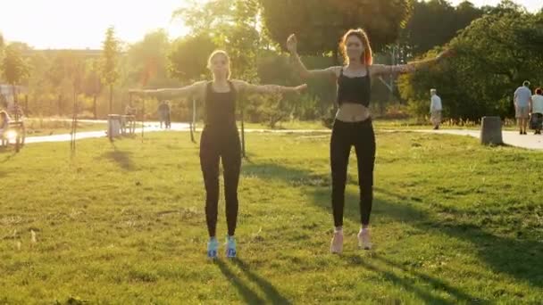 Две спортивные девушки тренируются вместе в солнечном парке. Занятия фитнесом, аэробикой и упражнениями на растяжку, здоровый и спортивный образ жизни — стоковое видео