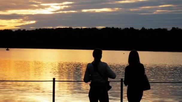 Silueta de primer plano de una turista, dos mujeres se quedan en un puente sobre el lago y disfrutan de la puesta de sol en la noche — Vídeo de stock