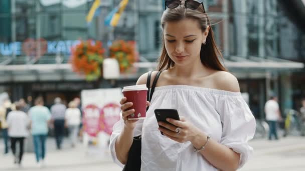 Sorridente donna carina con una tazza di caffè utilizzare il telefono cellulare sulla strada affollata della città — Video Stock