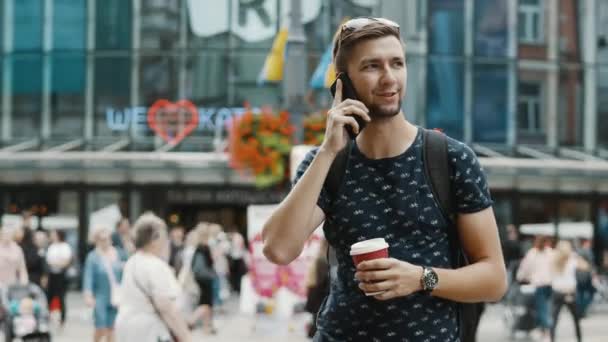 Buon turista maschio con una tazza di caffè parla dal telefono cellulare sulla strada affollata della città, con uno zaino, persone sullo sfondo — Video Stock