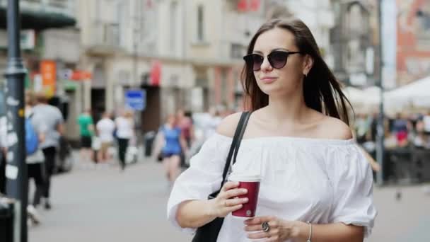 Boldog női turista egy csésze kávé áll a zsúfolt városi utcán sétál, és várja a srác a dátumot, az emberek és a bevásárló központ a háttérben — Stock videók