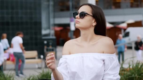 Feliz mujer de negocios descansar en el banco en la calle de la ciudad llena de gente, con gafas de sol, la gente de fondo — Vídeos de Stock