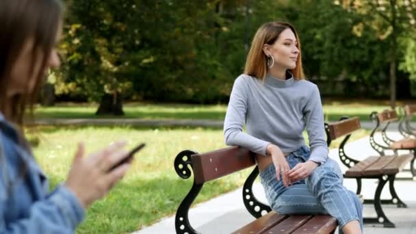 Hermosa pelirroja resto en el banco en el parque, en un borroso primer plano chica utiliza un teléfono móvil — Vídeos de Stock