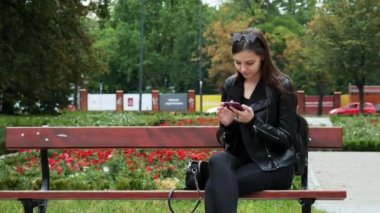 Neşeli genç kadın bir parkta bir bankta cep telefonu kullanarak, genç kadın internette gezinirken gülümseyen