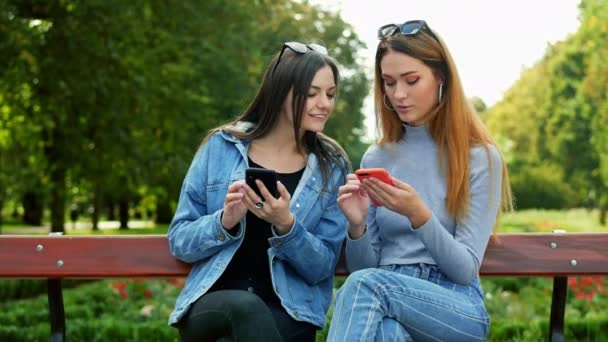 Dos amigas sentadas en un banco en el parque y usando el teléfono móvil, eligiendo fotos — Vídeos de Stock