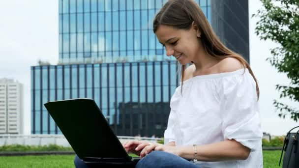 Nahaufnahme Porträt einer jungen Frau, die auf Gras sitzt und Laptop benutzt, Geschäftsfrau tippt auf Tastatur, modernes Geschäftsglasgebäude im Hintergrund — Stockvideo