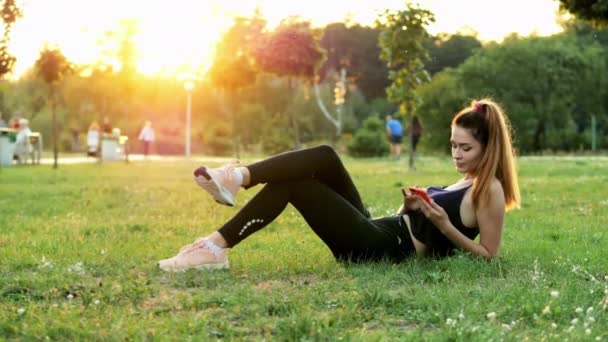 スポーティかわいい女性は、日没に市立公園の草の上に横たわっている間に携帯電話を使用し、トレーニング中に休憩時間を持っています — ストック動画