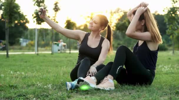 Dvě atletické ženy ve sportovním oblečení, které si berou na mobil, sedí na trávě v městském parku při západu slunce, mají při výcviku fotografickou relaci — Stock video