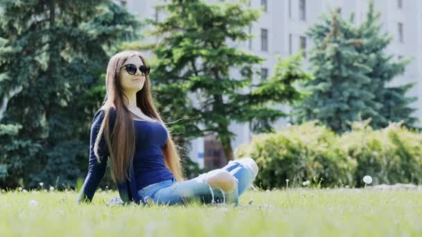 Heureuse femme repose sur l'herbe dans le parc de la ville, portant des lunettes de soleil, arbre sur le fond — Video