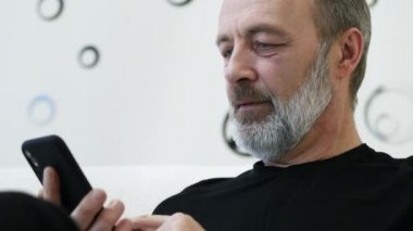 Gri sakallı orta yaşlı bir adam telefonunu evde kullanıyor, yakın plan.