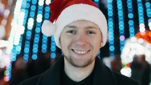 Junger glücklicher Mann Porträt mit Neujahrsmütze Lächeln und Blick in die Kamera, Bokeh von Weihnachtsgirlanden Lichter — Stockvideo