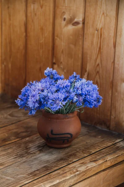 Flowers of cornflower in white earthenware. The vase is on a wooden shelf. A bouquet of flowers in a vase of clay. — Stock Photo, Image