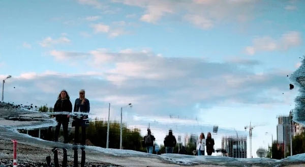 Şehir, insanlar ve gökyüzü bir su birikintisi yansıyan.