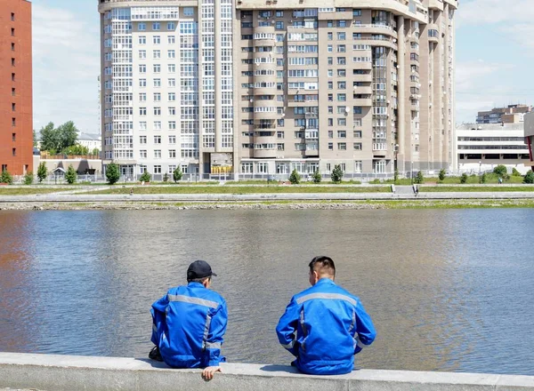 Çok katlı bir binanın arka planında şehir göletinin kıyısında oturan iş kıyafetleri içinde iki adam.