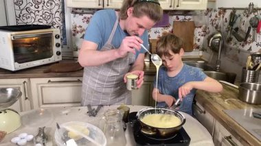 Önlüklü genç bir baba ve küçük oğlu mutfakta birlikte yemek pişiriyorlar. Aile tatillerini paylaşırlar, yemek hazırlama ve sıcak bir yuva ortamında eğlenceli etkileşimlerle eğlenirler.. 