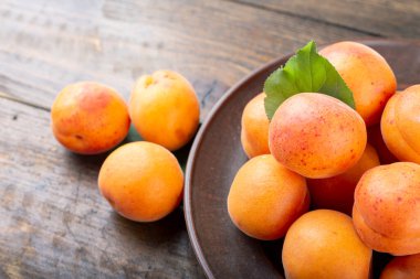 Ahşap bir zeminde, bir masada, kil bir tabakta olgun kayısılar. Kayısıların yanında yeşil izin.