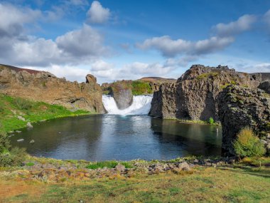 Güneşli bir günde Hjalparfoss şelalesi ve lagünün üzerinde uçan destansı hava aracı görüntüsü.