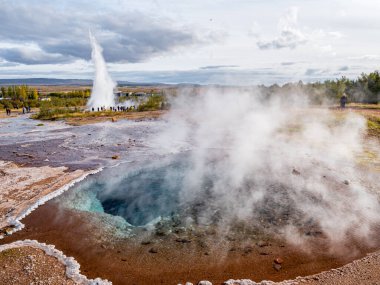 Arkasında Strokkur büyük gayzer buharı olan Haukadalur jeotermal bölgesinde buharlı mavi havuz. İzlanda 'nın en çok ziyaret edilen ve simgeleşen manzaralı yerlerinden biri.