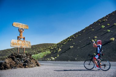 Lanzarote, İspanya. Şubat 2019 civarı. Bisikletçi, Lanzarote, Kanarya Adaları 'ndaki Timanfaya volkanlarının girişindeki tabelayı çekiyor.