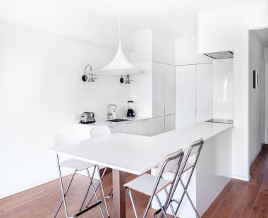 Beyaz dolaplı, şık, parlak bir mutfak. Ahşap zemin, beyaz masa, endüstriyel sandalyeler ve büyük tavan penceresi olan geniş modern iç mekan..