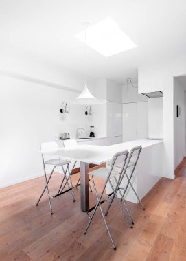 Beyaz dolaplı, şık, parlak bir mutfak. Ahşap zemin, beyaz masa, endüstriyel sandalyeler ve büyük tavan penceresi olan geniş modern iç mekan..