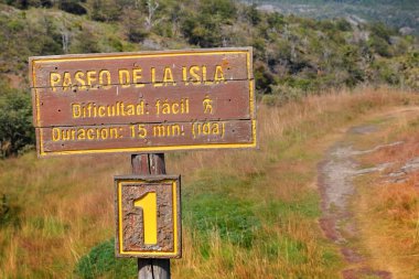 Yeşil, kahverengi ve turuncu bitki örtüsü ile çevrili Tierra del Fuego Milli Parkı'ndaki Paseo de la Isla yürüyüş parkurunu gösteren ahşap tabela.