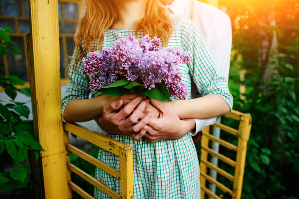 Man hugging his girlfriend who is holding a bouquet of lilacs.