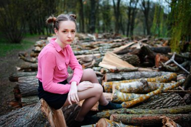 Beautiful young woman sitting on stack of felled tree trunks in the forest. clipart