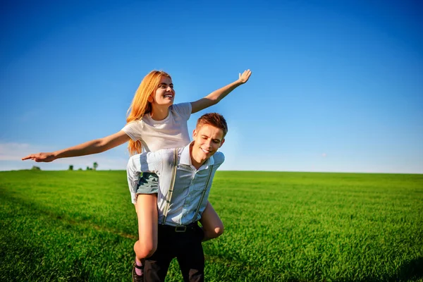 彼の戻って幸せの女性は 彼女の腕を引っ張るし 青い空と緑のフィールドの背景に飛行をシミュレートに保持している男の笑みを浮かべて — ストック写真
