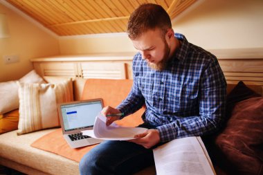 Genç adam eve bir dizüstü bilgisayarda çalışırken belgeleri tutan bir kanepede oturan eğitim.