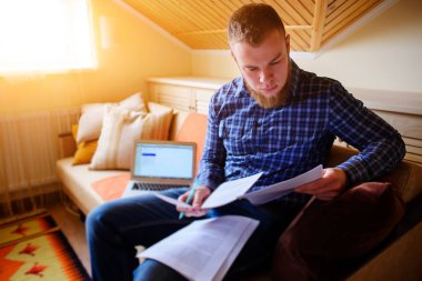 Genç adam eve bir dizüstü bilgisayarda çalışırken belgeleri tutan bir kanepede oturan eğitim.